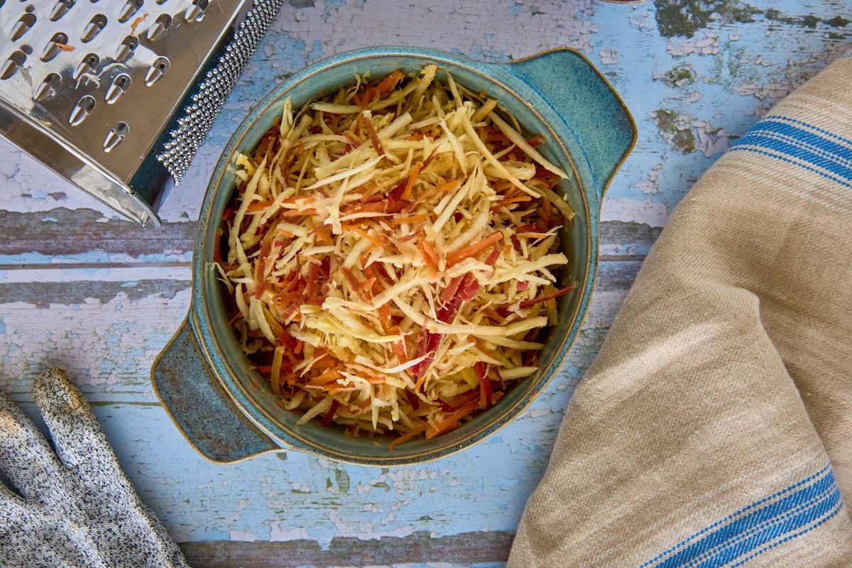 grated carrots and parsnips in a bowl with a kitchen towel, box grater, and grating glove