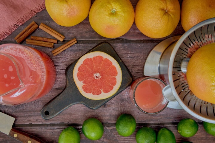 grapefruits, limes, a halved grapefruit in a citrus juicer, a halved grapefruit on a cutting board, a knife, a glass collecting juice, a pitcher of juice, a napkin, and cinnamon sticks