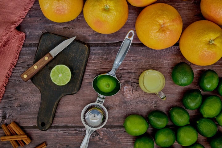 grapefruits, limes, a halved lime in a citrus squeezer, a halved lime and knife on a cutting board, a small pitcher of lime juice, cinnamon sticks, and a napkin