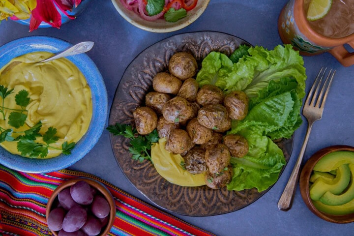 huancaína sauce in a bowl, surrounded by a piece of fabric, olives, a plate of lettuce, roasted baby potatoes with huancaína sauce, avocado slices, a mug of chirulín cocktail, a vase of flowers, avocado slices, and pickled onions and chilis