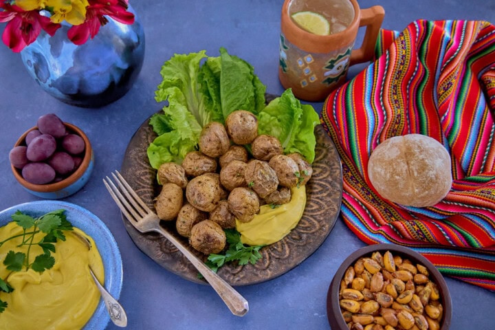 huancaína sauce in a bowl, surrounded by a piece of fabric, olives, a plate of lettuce, roasted baby potatoes with huancaína sauce, a bread roll, a mug of chirulín cocktail, a vase of flowers, and toasted corn