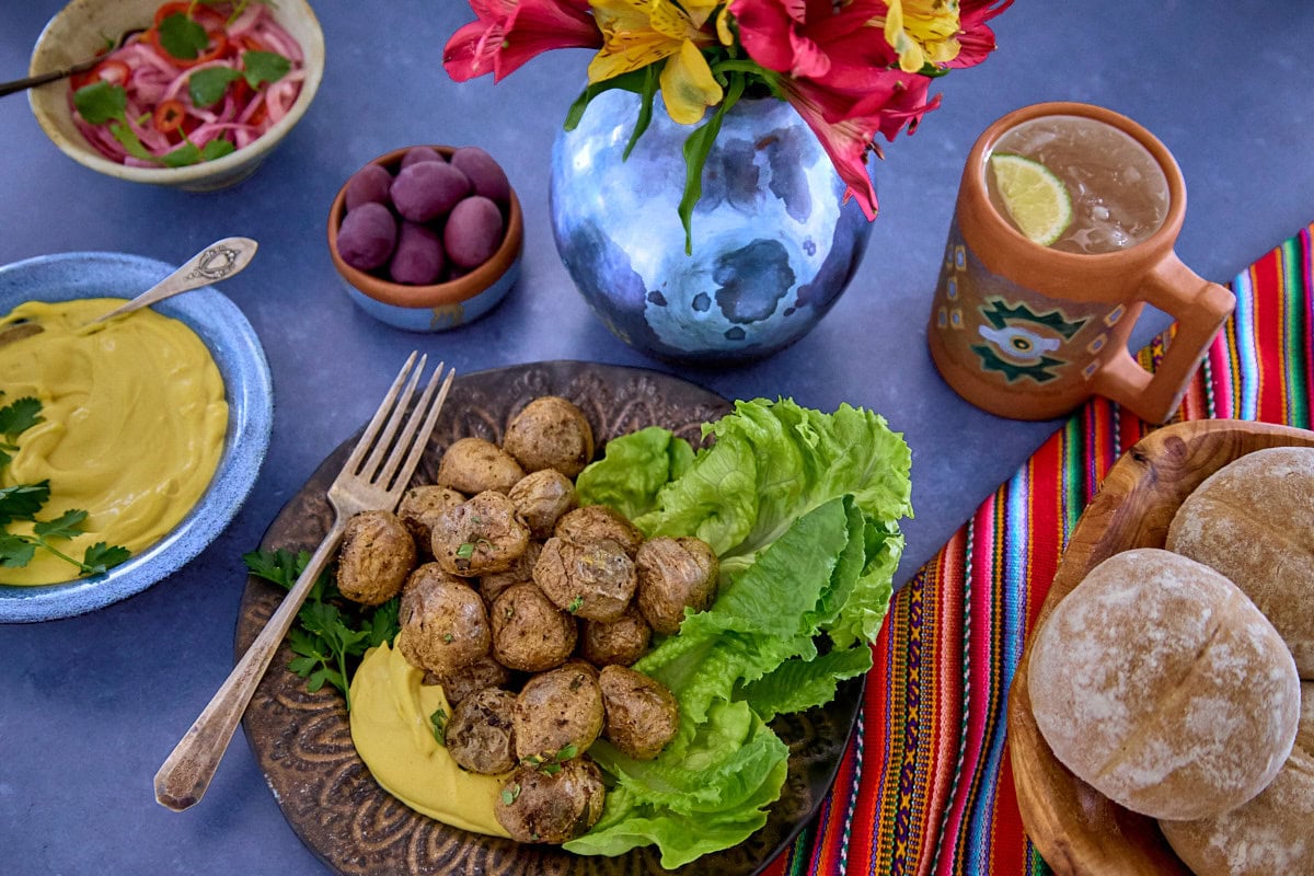 huancaína sauce in a bowl, surrounded by a piece of fabric, a plate of lettuce, roasted baby potatoes with huancaína sauce, olives, a mug of chirulín cocktail, a vase of flowers, bread rolls, and pickled onions and chilis