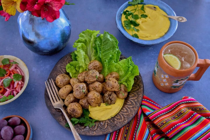 huancaína sauce in a bowl, surrounded by a piece of fabric, a plate of lettuce, roasted baby potatoes with huancaína sauce, olives, a mug of chirulín cocktail, a vase of flowers, and pickled onions and chilis