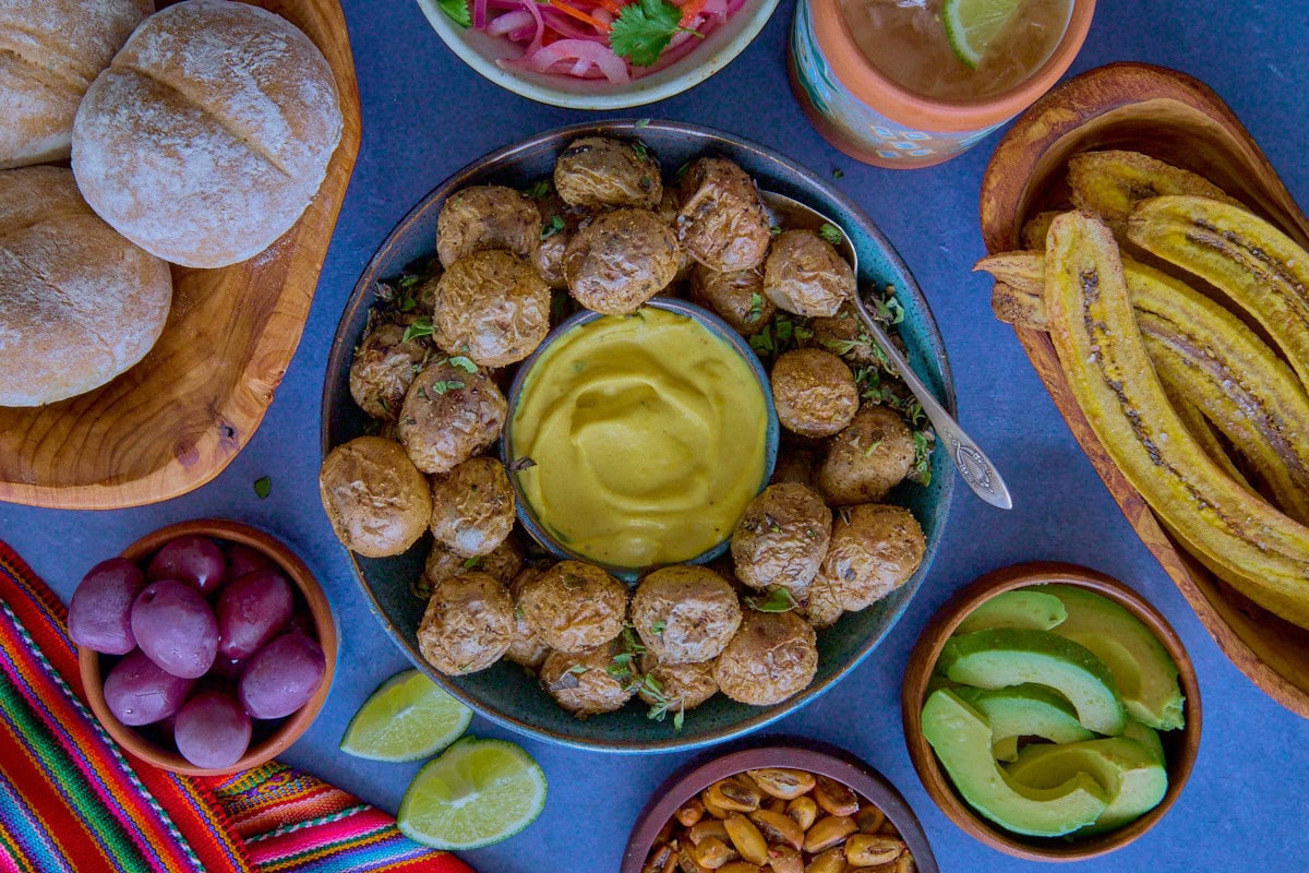 huancaína sauce and potatoes in a bowl, surrounded by a piece of fabric, toasted plantain chips, avocado slices, bread rolls, pickled onions and chilis, and toasted corn