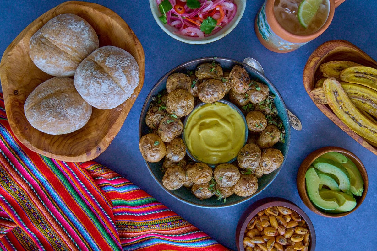 huancaína sauce and potatoes in a bowl, surrounded by a piece of fabric, toasted plantain chips, avocado slices, bread rolls, pickled onions and chilis, and toasted corn