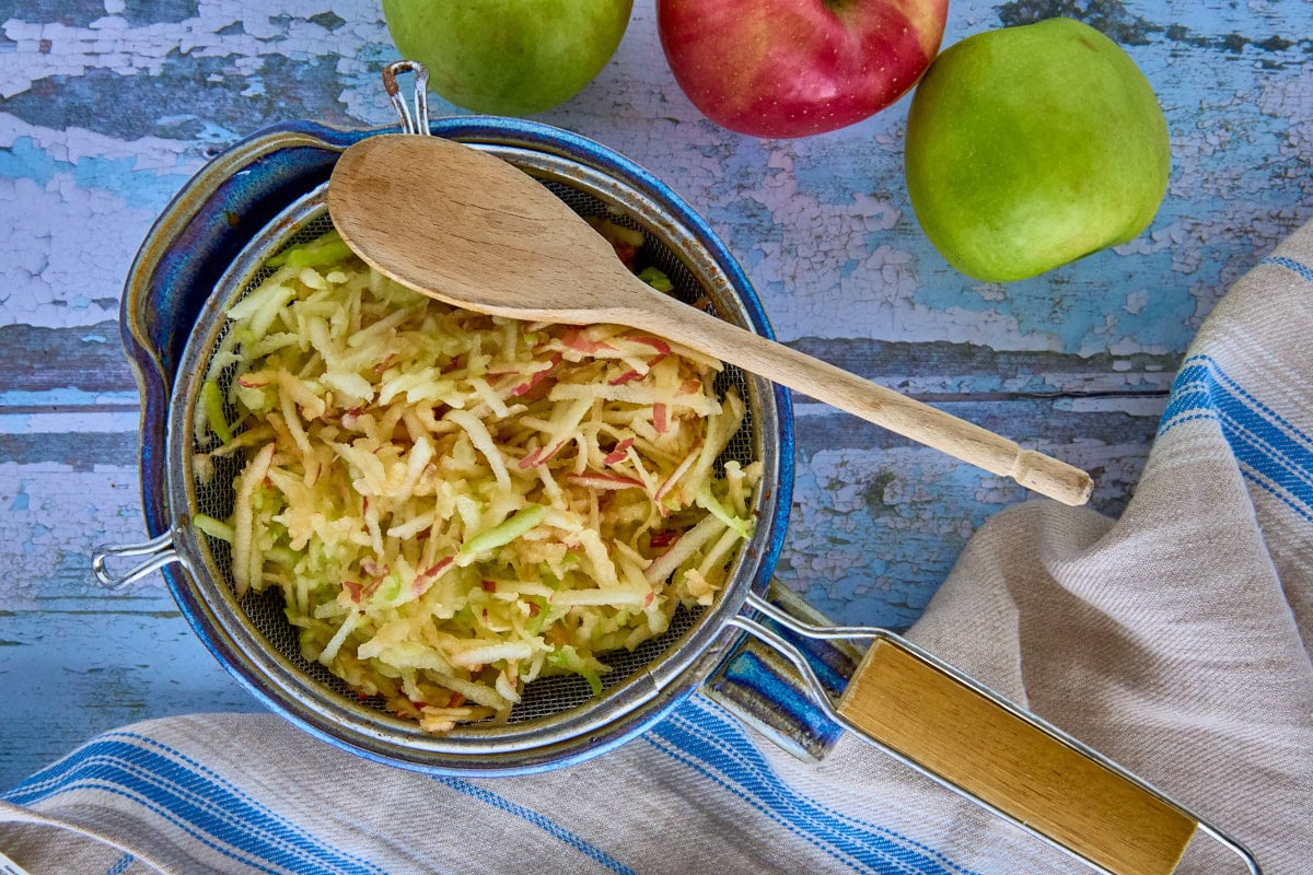 grated apples in a sieve over a mixing bowl with a wooden spoon, kitchen towel, and apples
