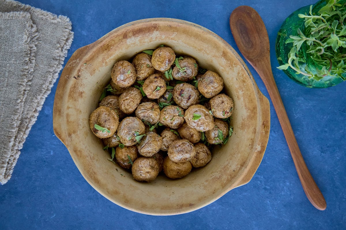 seasoned roasted potatoes in a bowl with a napkin, wooden mixing spoon, and vase of oregano