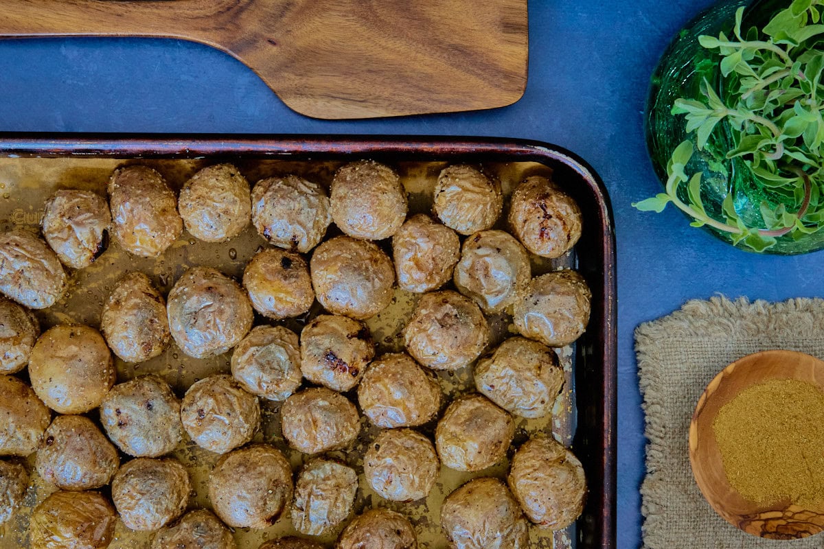 roasted potatoes in a sheet pan, a napkin, bowl of cumin, vase of oregano, and wooden spatula