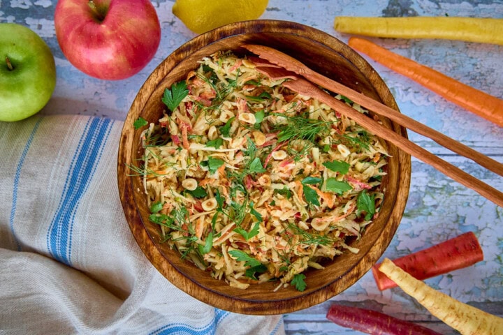 a bowl of salad with serving utensils, kitchen towel, apples, carrots, and parsnip
