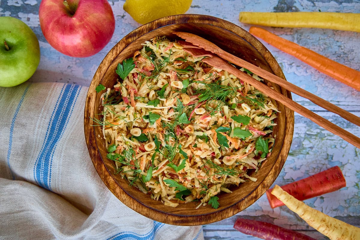 a bowl of salad with serving utensils, kitchen towel, apples, carrots, and parsnip