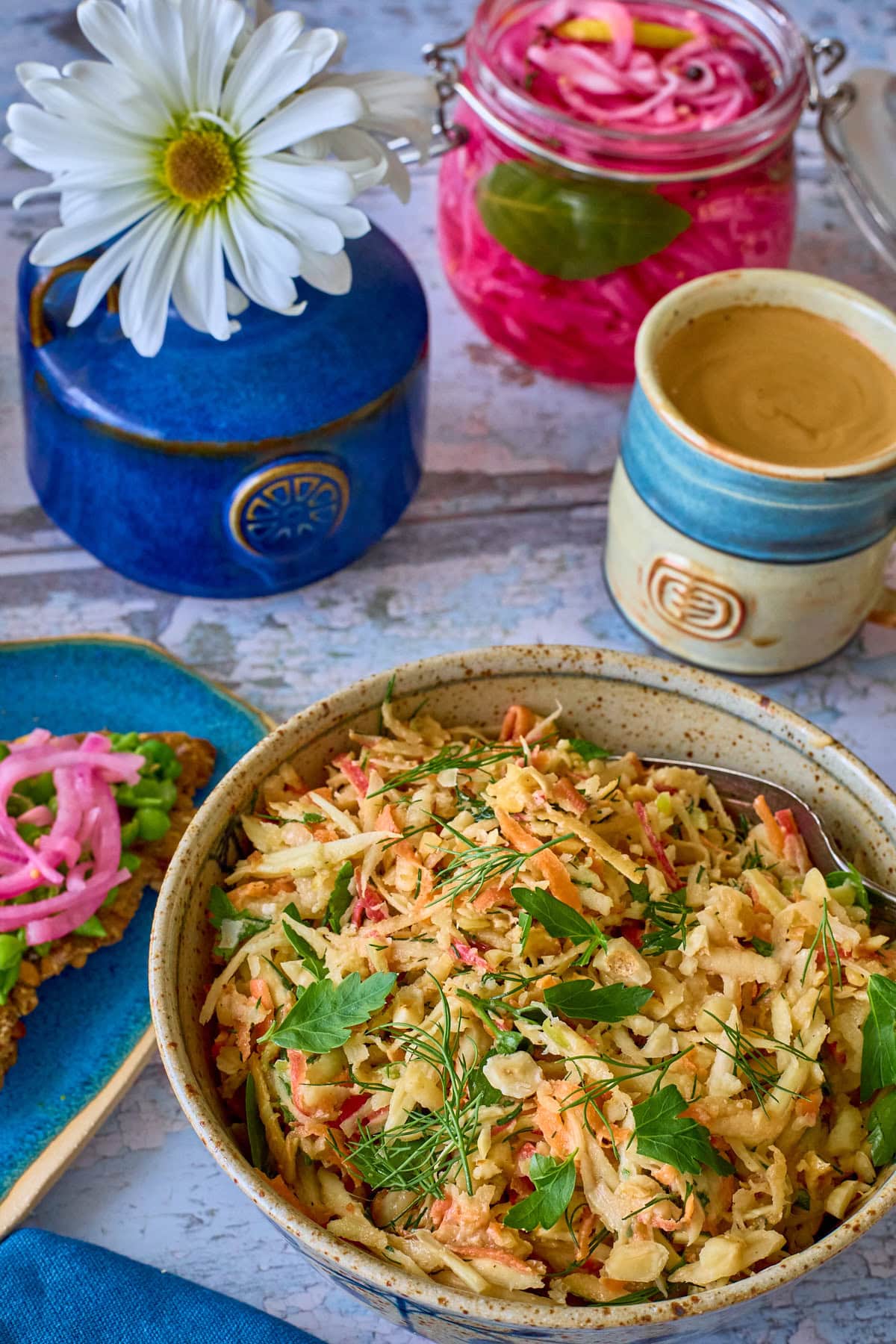 a bowl of parsnip salad with a fork, plate with crispbread topped with peas and onions, napkin, vase of daisies, jar of pickled onions, and cup of coffee