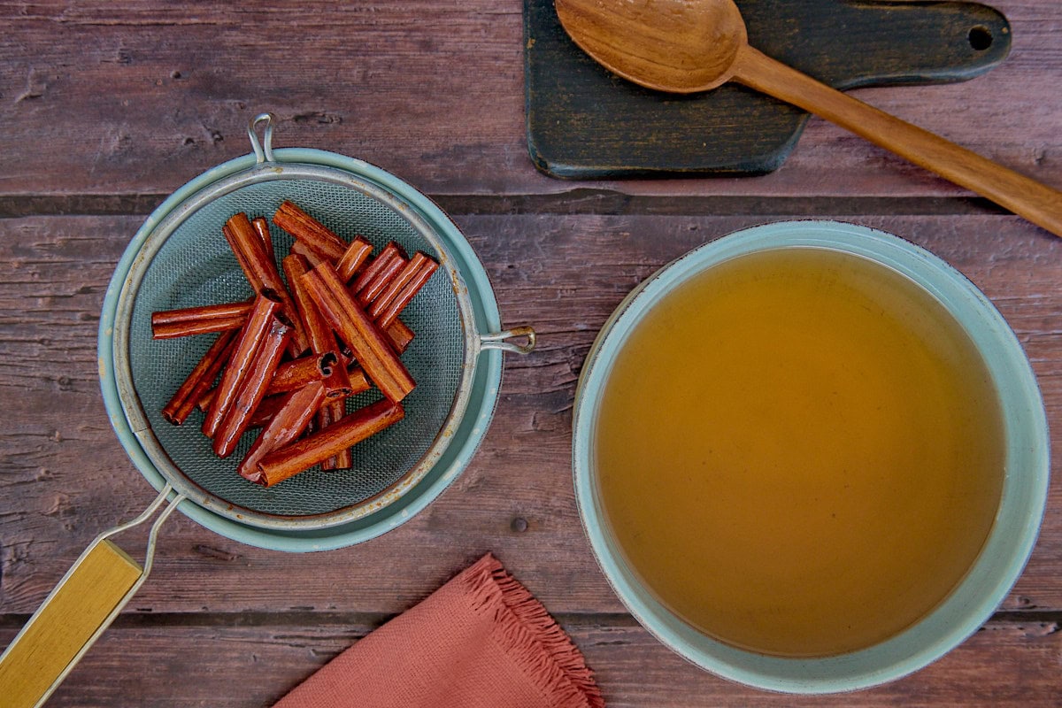 cinnamon sticks in a sieve over a bowl, a wooden mixing spoon on a cutting board, a bowl of cinnamon syrup, and a napkin