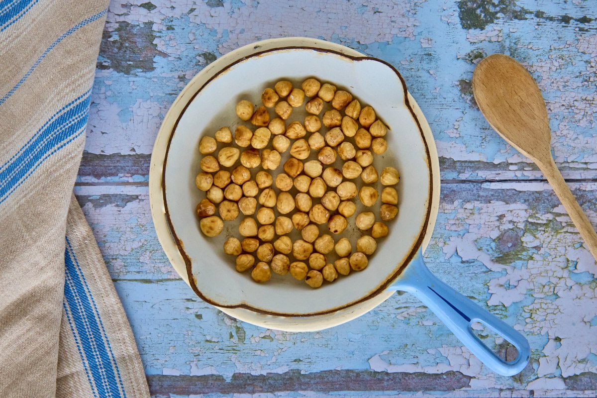 a skillet with toasted hazelnuts on a trivet, with a kitchen towel and wooden mixing spoon