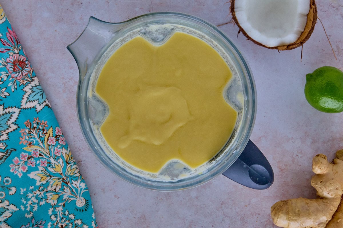 a blended cocktail in a blender with a kitchen towel, halved coconut, lime, and ginger root in the background