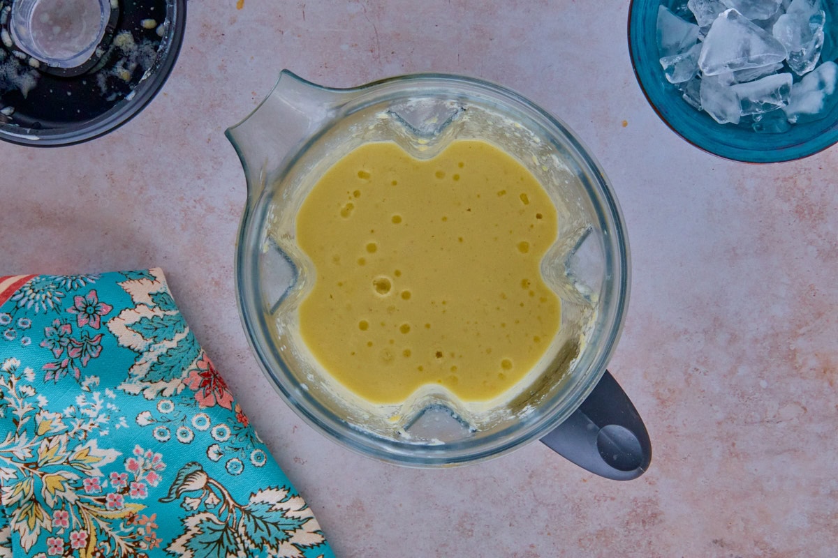 blended drink ingredients in a blender with a kitchen towel, blender top, and bowl of ice
