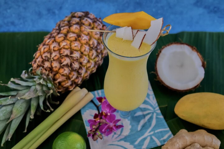 a cocktail in a glass on banana leaves on a cocktail napkin with a pineapple, halved coconut, lemongrass stalks, orchid flowers, a lime, a ginger root, and a mango in the background