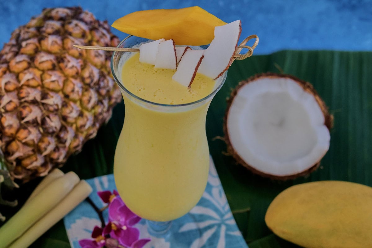 a cocktail in a glass on banana leaves on a cocktail napkin with a pineapple, halved coconut, lemongrass stalks, orchid flowers, and a mango in the background
