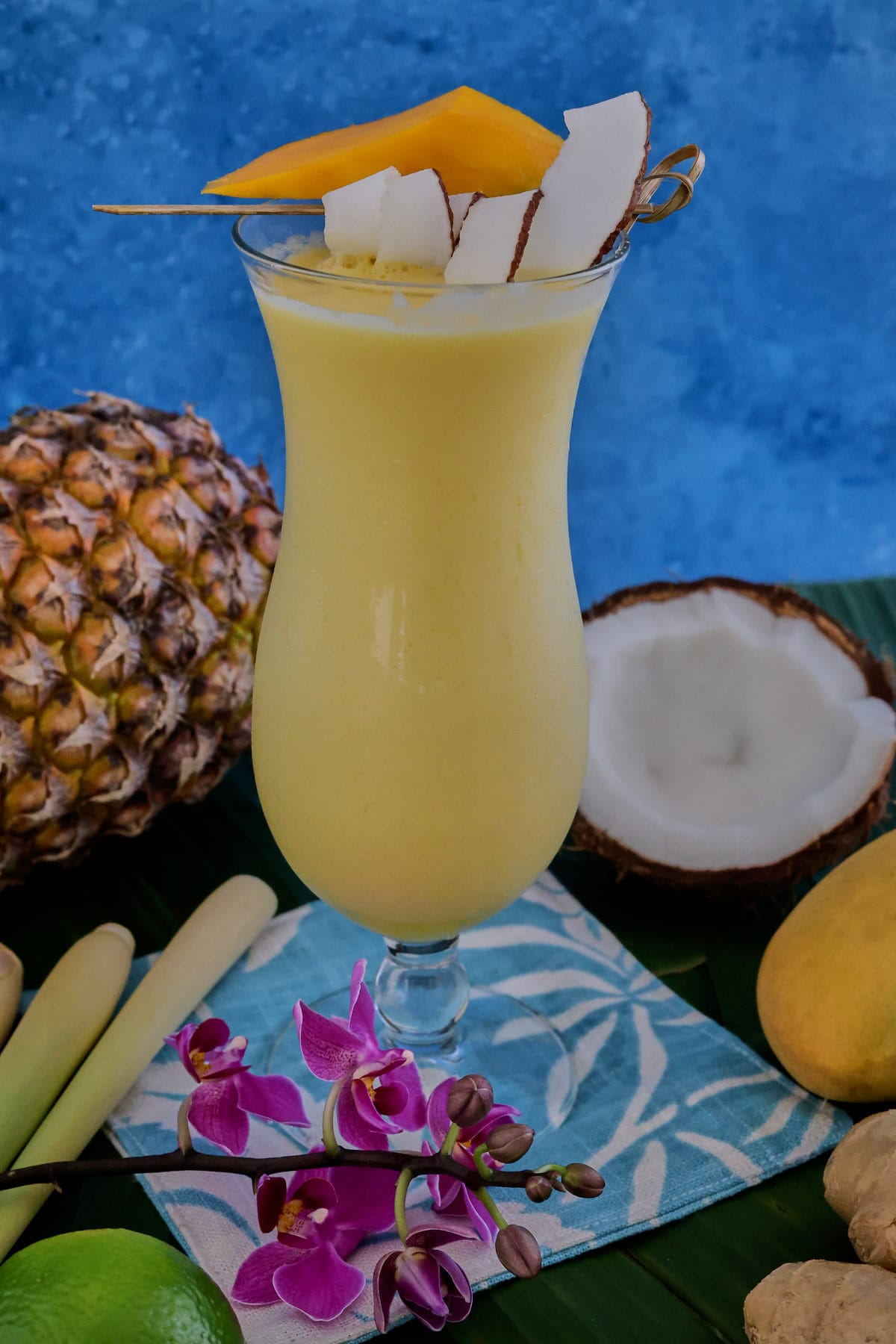 a cocktail in a glass on banana leaves on a cocktail napkin with a pineapple, halved coconut, lemongrass stalks, orchid flowers, and a mango in the background