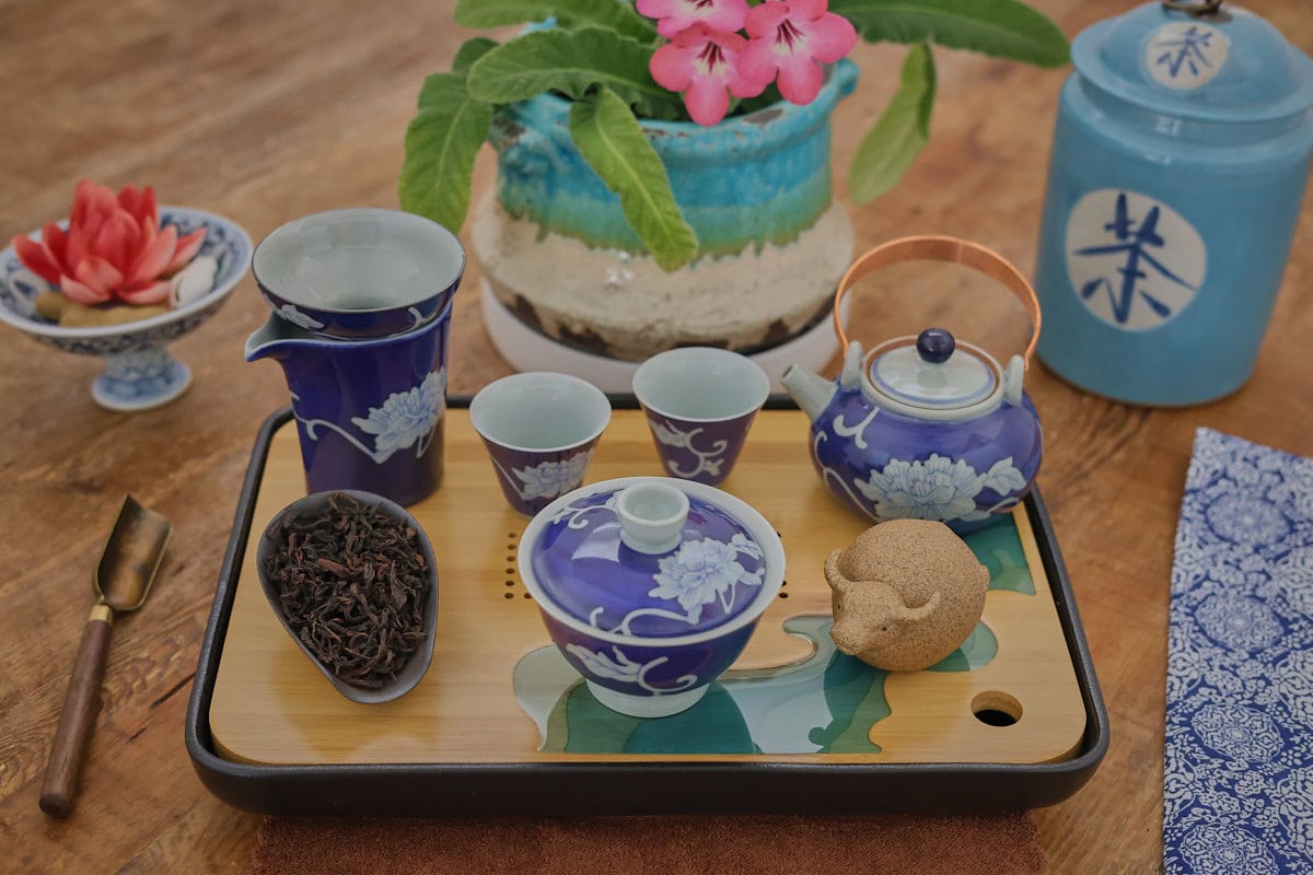 a ceramic gong fu tea brewing set with teaware, dried tea, a brewing tray, scoop, footed bowl of tea pets, a flowering plant, tea storage jar, and napkin