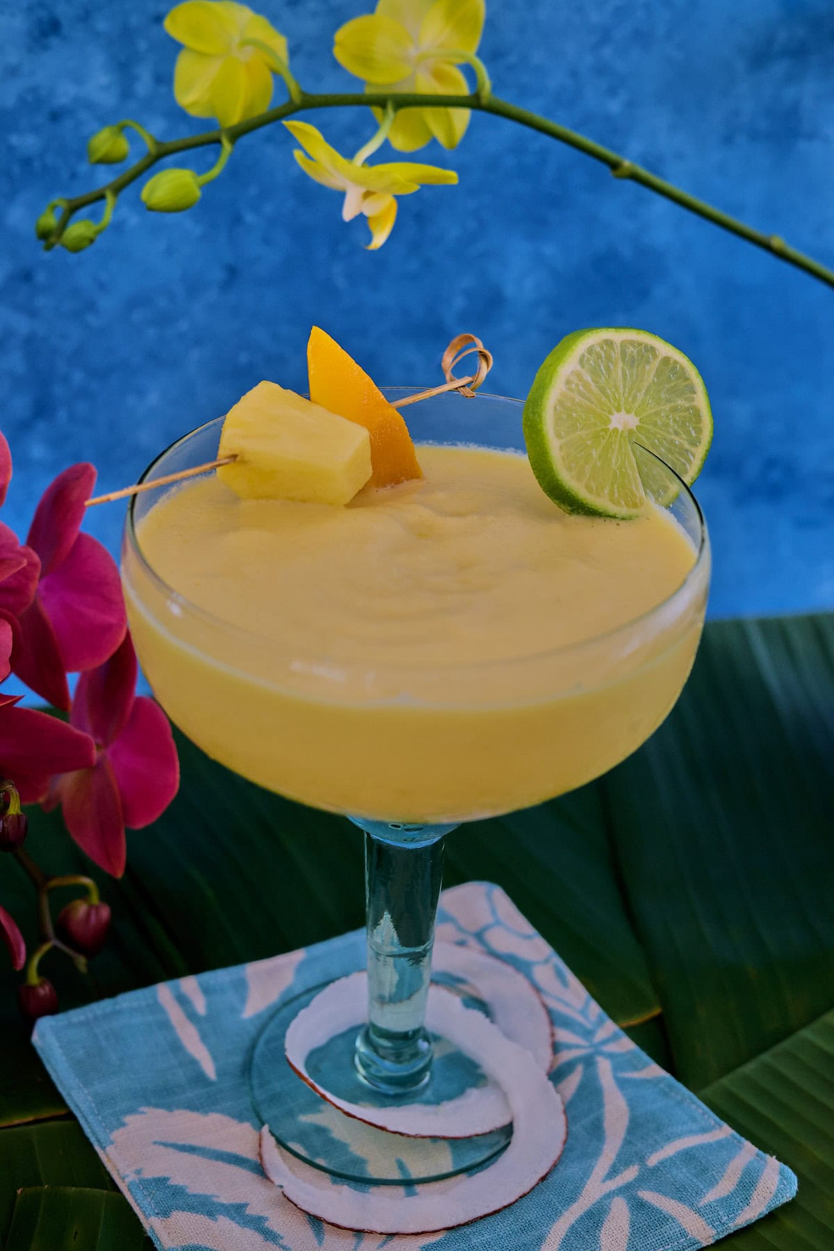 a cocktail in a glass on banana leaves on a cocktail napkin with a strip of fresh coconut wrapped around the glass base and orchid flowers in the background