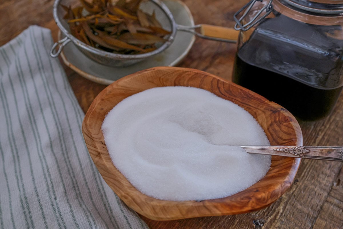 a wooden bowl with sugar and a spoon, a kitchen towel, a strainer full of steeped lemon verbena leaves on a plate, and a jar of liqueur on a table