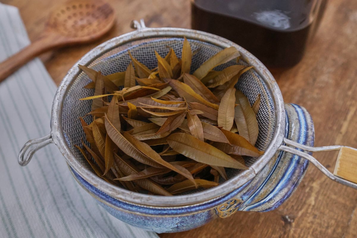 steeped lemon verbena leaves in a strainer over a bowl, with a kitchen towel, wood straining spoon, and jar of liqueur in the background