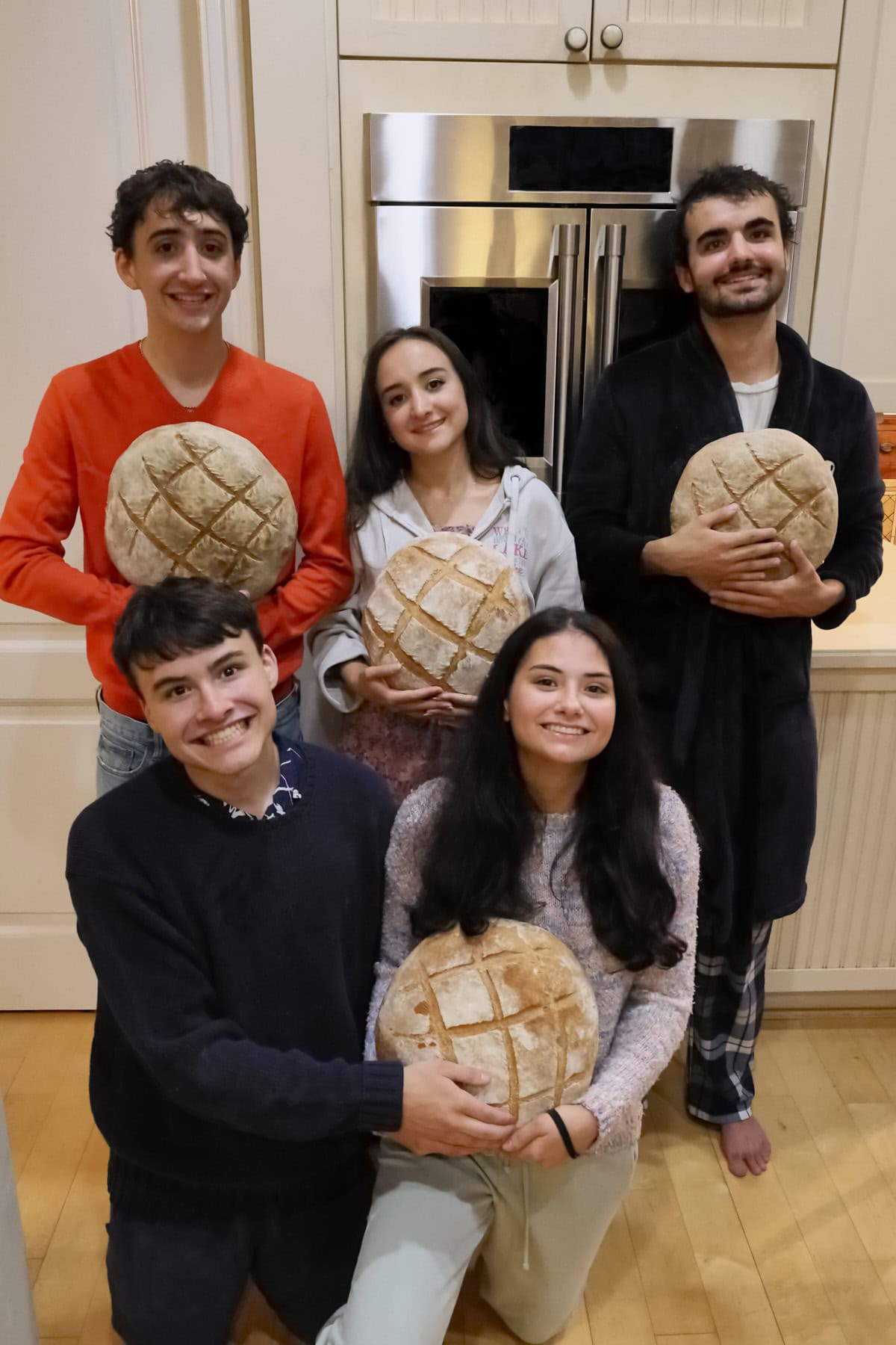 5 young adults in front of double wall ovens, holding 4 large round loaves of homemade bread