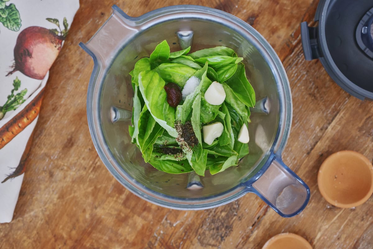 a blender container with ingredients for green basil vinaigrette, with a blender lid, kitchen towel, and 2 small bowls in the background