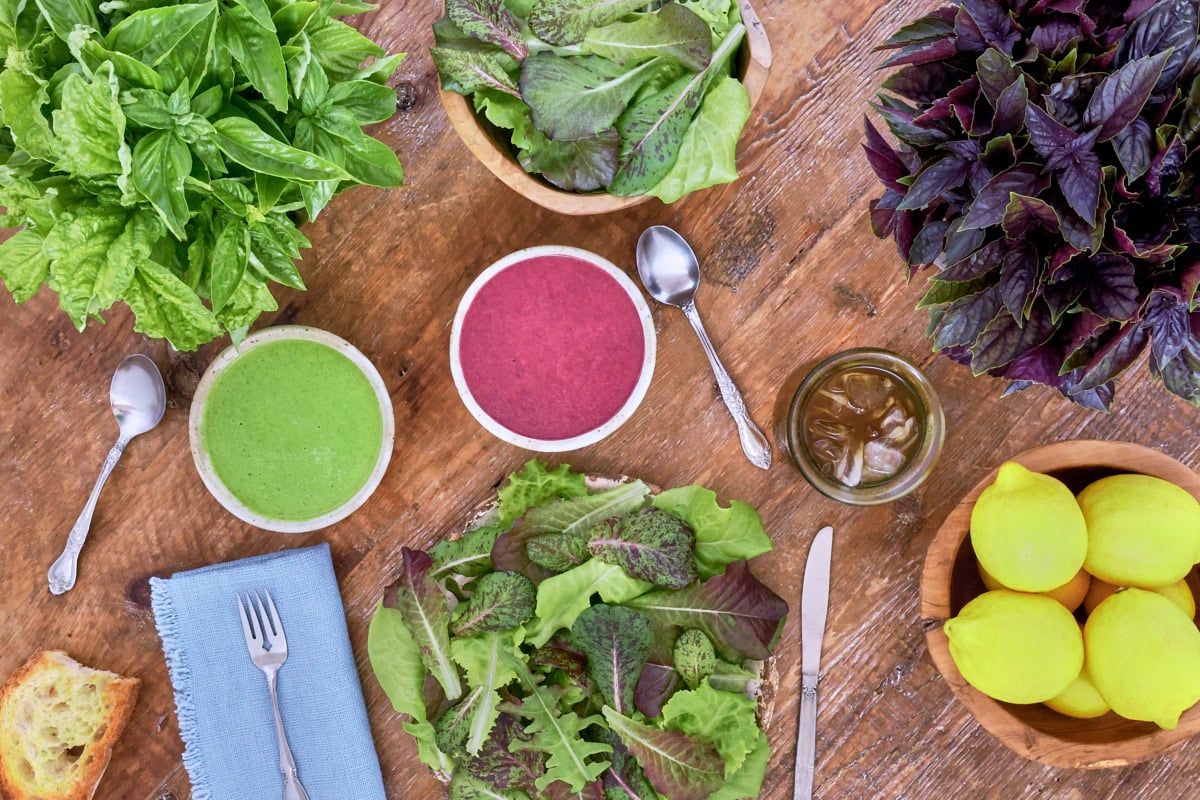 a plate of salad, knife, fork, napkin, piece of toasted bread, bowl of lemons, bowls of green and purple basil vinaigrette, spoons, glass of iced tea, bowl of lettuce, and bowls of green and purple basil leaves