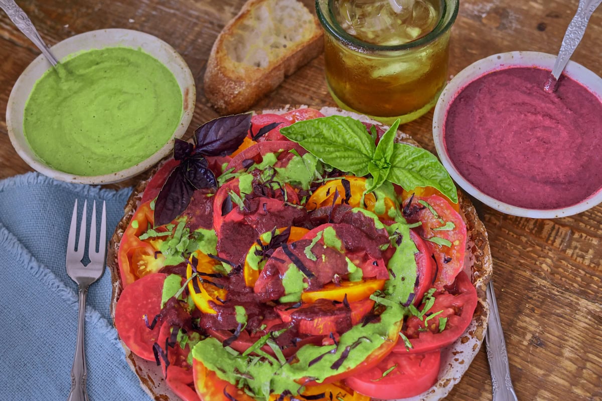 a plate of sliced tomatoes with vinaigrette and sliced basil on top, with a knife, fork, napkin, bowls of green and purple basil vinaigrette, spoons, a piece of toasted bread, and a glass of iced tea