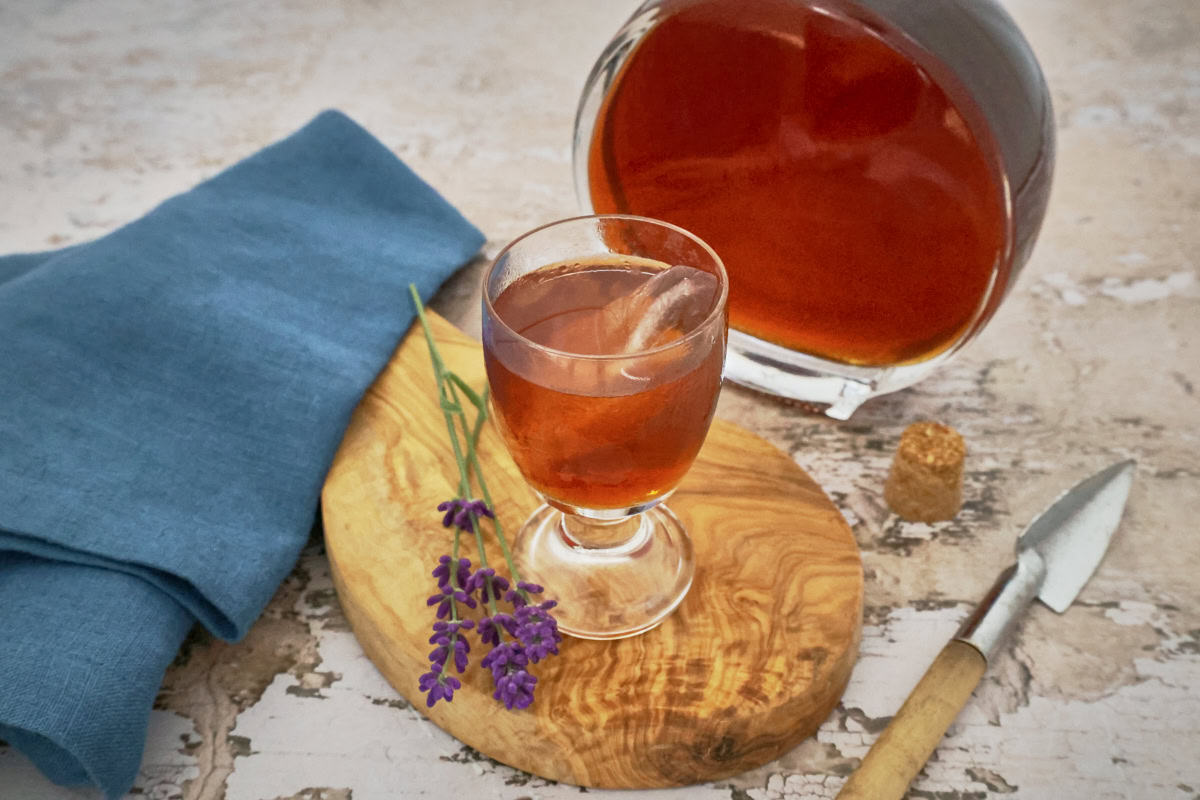 a glass and bottle of lavender liqueur, lavender stems, napkin, wooden tray, lemon, vanilla beans, and mixing spoon