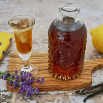 a glass and bottle of lavender liqueur, lavender stems, napkin, wooden tray, lemon, and vanilla beans