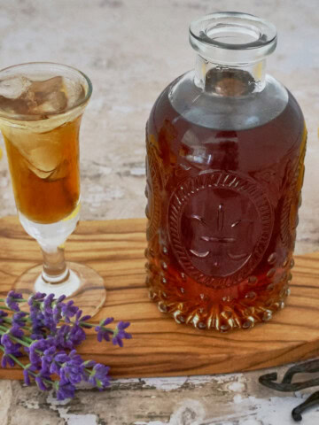 a glass and bottle of lavender liqueur, lavender stems, napkin, wooden tray, lemon, and vanilla beans