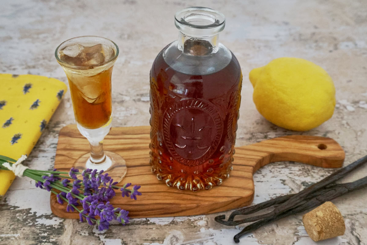 a glass and bottle of lavender liqueur, lavender stems, napkin, wooden tray, lemon, vanilla beans, and cork