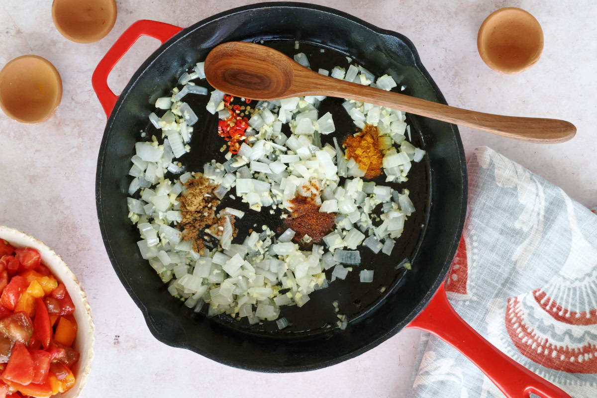 onions, garlic, ginger, and spices in a skillet with a wooden spoon, a kitchen towel, bowl of chopped tomatoes, and three small empty pinch bowls