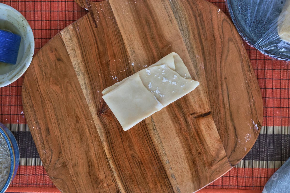 a partially-folded piece of dough on a wooden board, bowl of oil with a kitchen brush, bowl of flour, plate of dough pieces with plastic wrap, kitchen towel, and table runner
