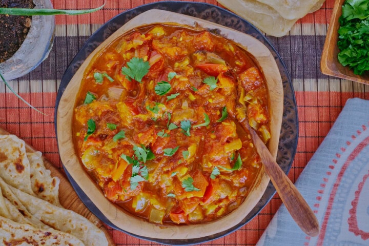 rougail sauce in a wooden bowl with a wooden spoon on a plate, with a table runner, kitchen towel, wooden board with farata flatbreads, more flatbreads above the bowl, and a plant