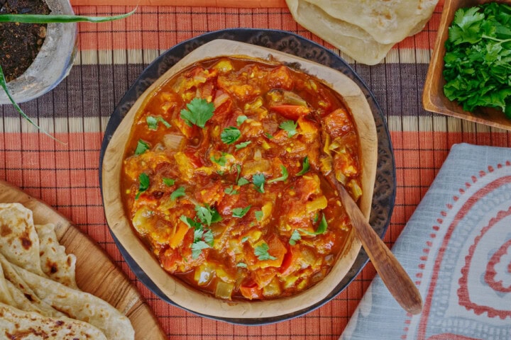 rougail sauce in a wooden bowl with a wooden spoon on a plate, with a table runner, kitchen towel, wooden board with farata flatbreads, more flatbreads above the bowl, and a plant