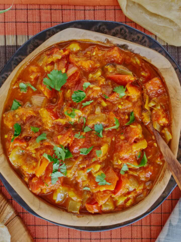 rougail sauce in a wooden bowl with a wooden spoon on a plate, with a table runner, kitchen towel, wooden board with farata flatbreads, more flatbreads above the bowl, and a plant