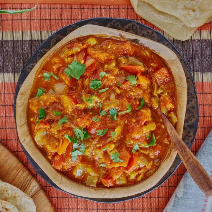 rougail sauce in a wooden bowl with a wooden spoon on a plate, with a table runner, kitchen towel, wooden board with farata flatbreads, more flatbreads above the bowl, and a plant