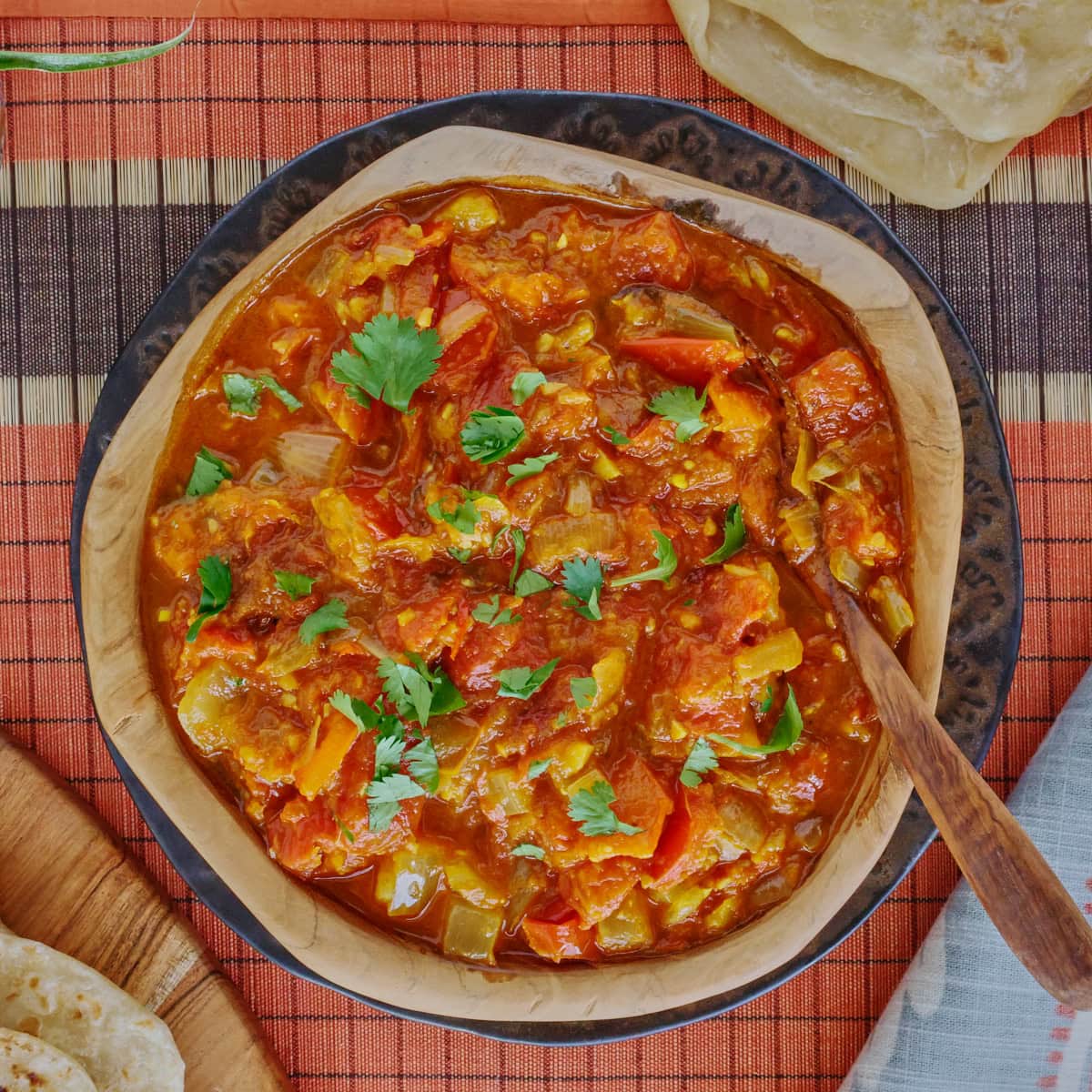 rougail sauce in a wooden bowl with a wooden spoon on a plate, with a table runner, kitchen towel, wooden board with farata flatbreads, more flatbreads above the bowl, and a plant