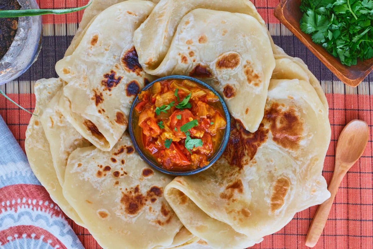 rougail sauce in a bowl surrounded by flatbreads on a table runner with a kitchen towel, wooden spoon, plant in a pot, and bowl of cilantro