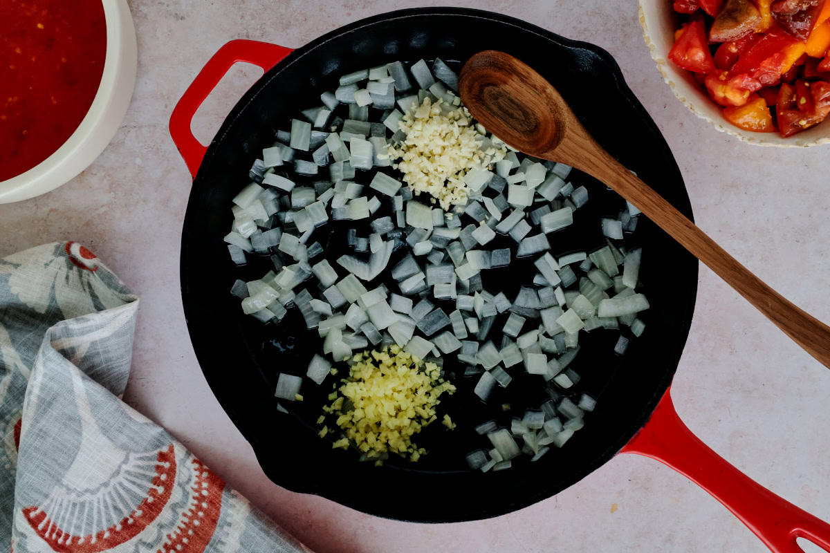onions, garlic, and ginger in a skillet with a wooden spoon, with a kitchen towel, bowl of tomato juice, and bowl of chopped tomatoes