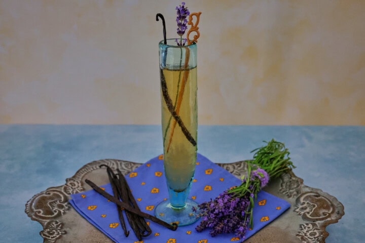 a cocktail in a champagne glass with a napkin, vanilla beans, and fresh lavender sprigs on a metal tray
