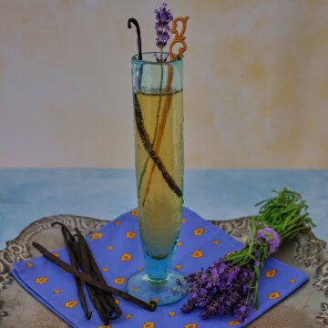 a cocktail in a champagne glass with a napkin, vanilla beans, and fresh lavender sprigs on a metal tray