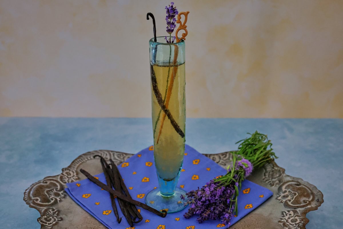 a cocktail in a champagne glass with a napkin, vanilla beans, and fresh lavender sprigs on a metal tray