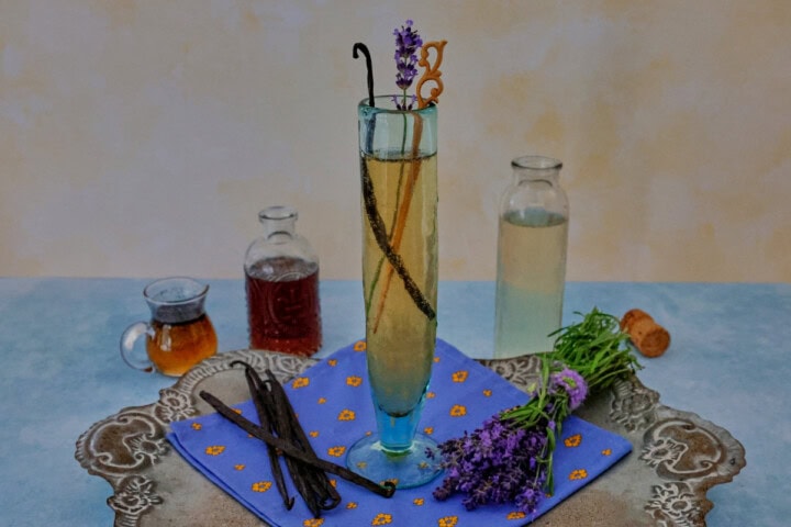 a cocktail in a champagne glass with a napkin, vanilla beans, and fresh lavender sprigs on a metal tray with a small pitcher of vanilla bean syrup, small bottle of lavender liqueur, and bottle of champagne in the background