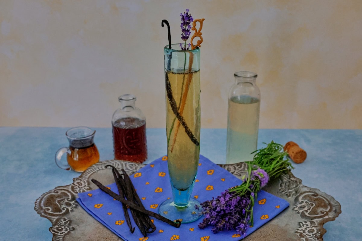 a cocktail in a champagne glass with a napkin, vanilla beans, and fresh lavender sprigs on a metal tray with a small pitcher of vanilla bean syrup, small bottle of lavender liqueur, and bottle of champagne in the background