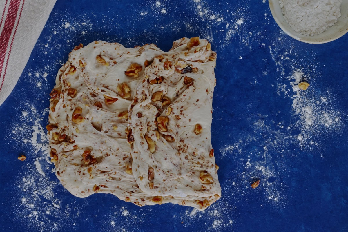 bread dough being folded like a letter, with a kitchen towel and bowl of flour alongside