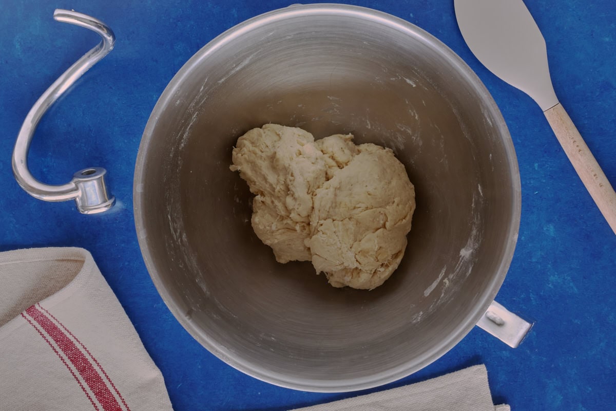 bread dough in a stand mixer, with a kitchen towel, dough hook, and spatula alongside